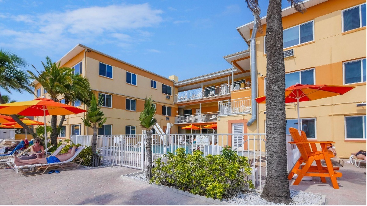 A sunny exterior view of a hotel with a fenced pool area, lounge chairs, and palm trees.
