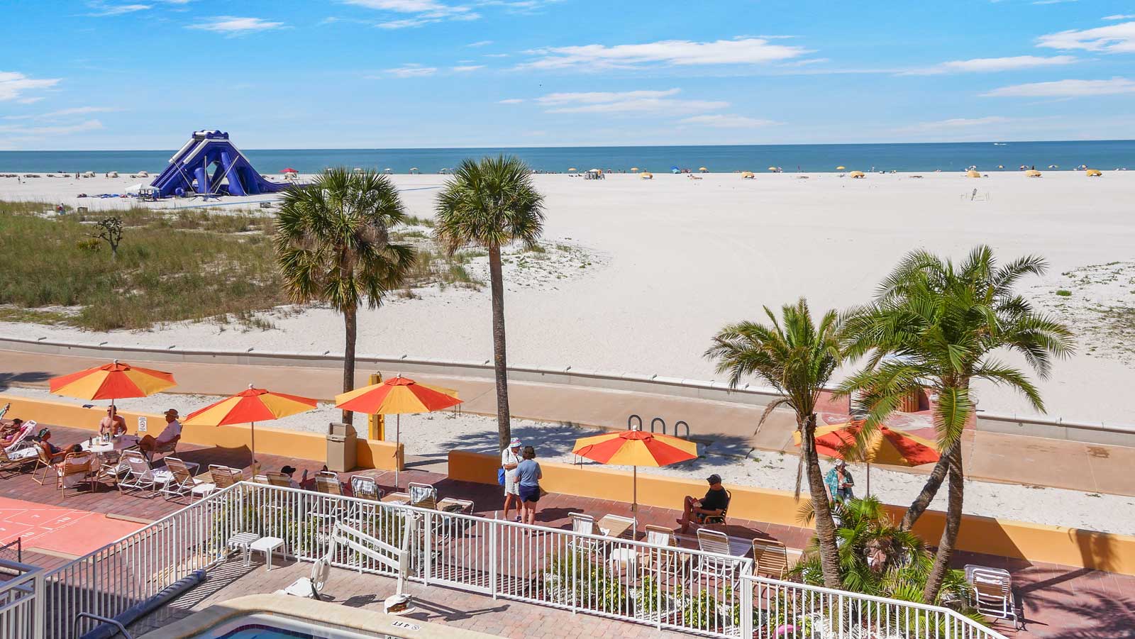 A beach view with palm trees, orange umbrellas, and a pool area in the foreground.